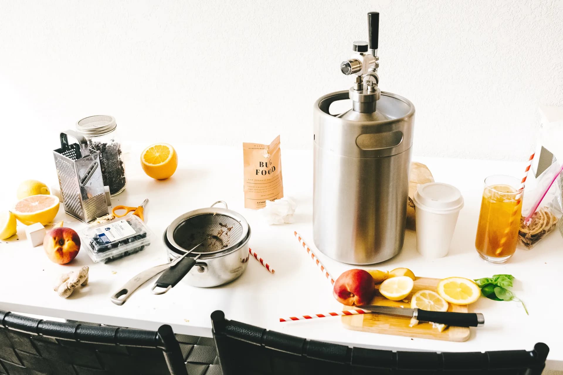 Sandpoint Soda brewing setup on a kitchen table - a stainless keg, Bug Food packet, box grater with fresh ginger, sliced citrus, berries, and a glass of soda